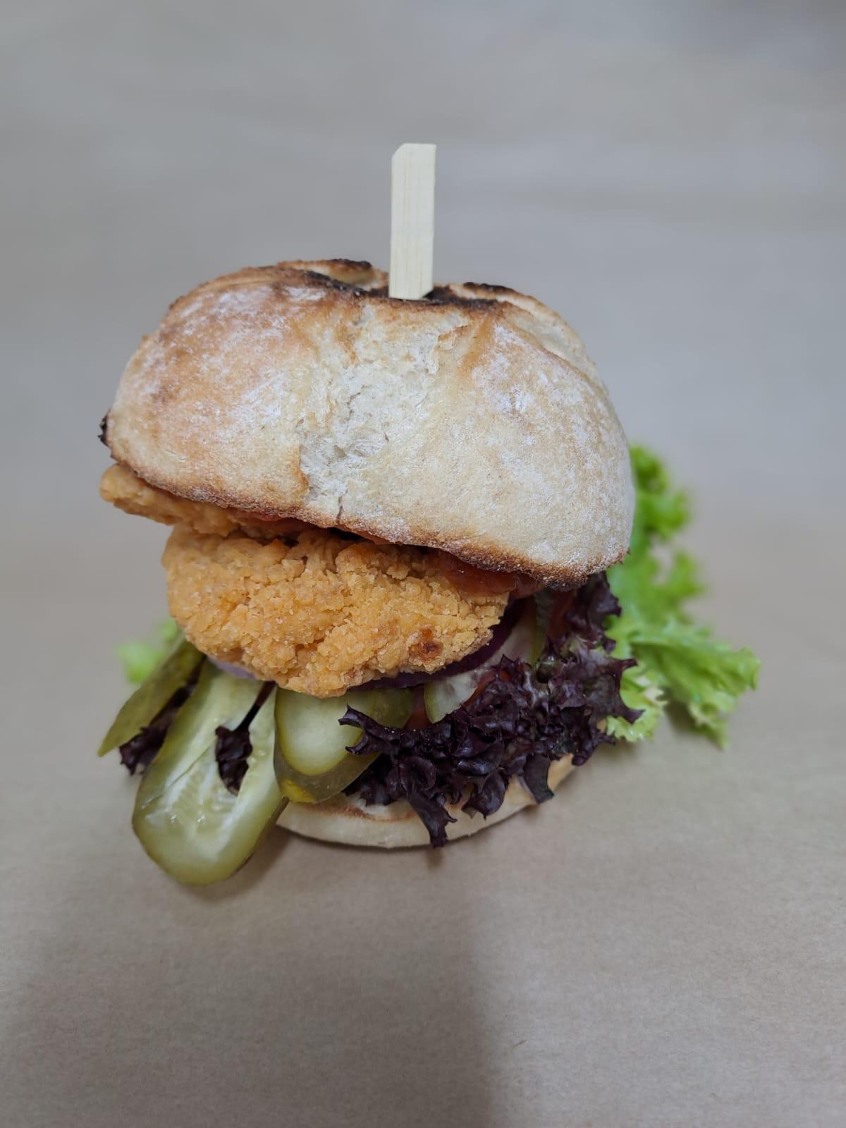 Burger z panierowanym kotletem, sałatą, piklami i czerwoną cebulą, spięty drewnianym patyczkiem, na neutralnym tle.