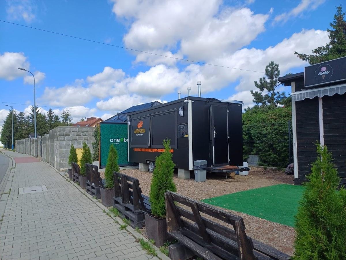 Czarny food truck z napisem 'Gastro Special' zaparkowany obok małego budynku z szyldem 'am', otoczone roślinami i ławkami, w tle błękitne niebo z chmurami.