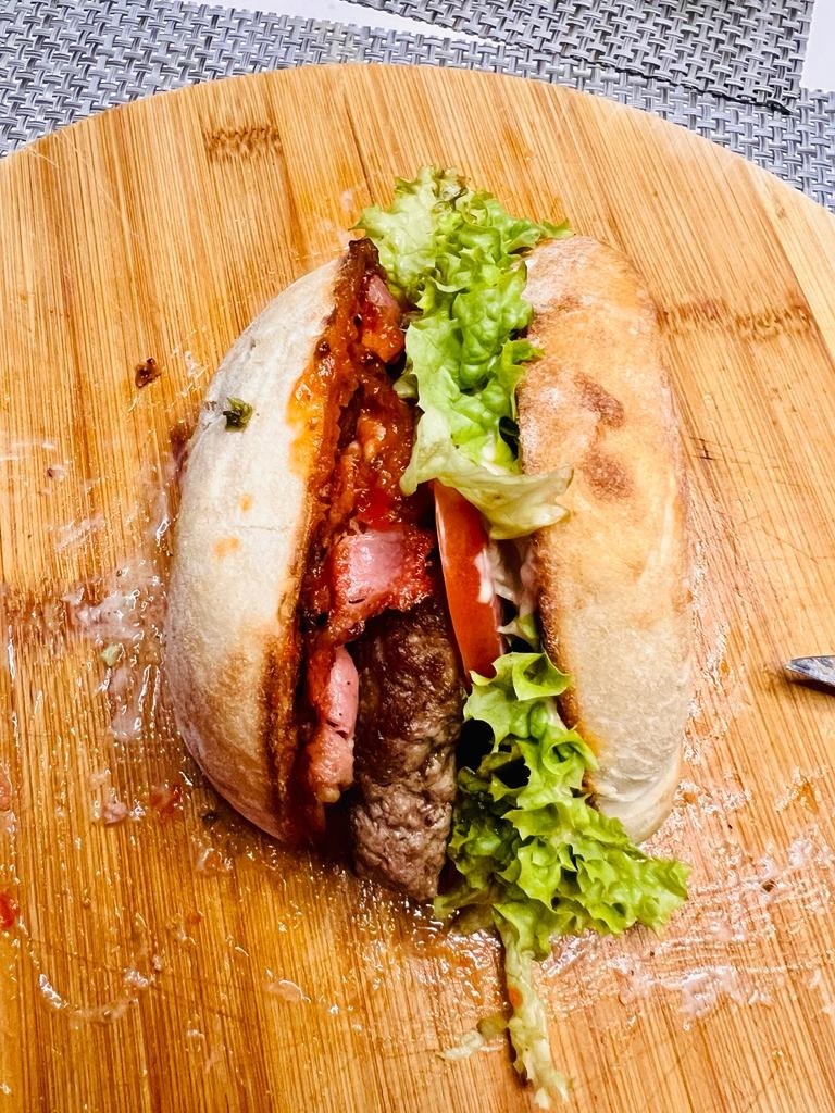 Duży hamburger z wołowiną, bekonem, pomidorem i sałatą w bułce ciabatta, podany na drewnianej desce do krojenia z widocznymi śladami użytkowania.