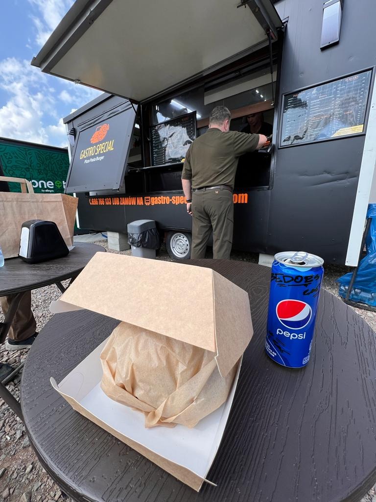 Burger zawinięty w papier w otwartym kartonowym pudełku na okrągłym stole, obok puszka Pepsi z graffiti, w tle czarny food truck z menu i klientem zamawiającym.