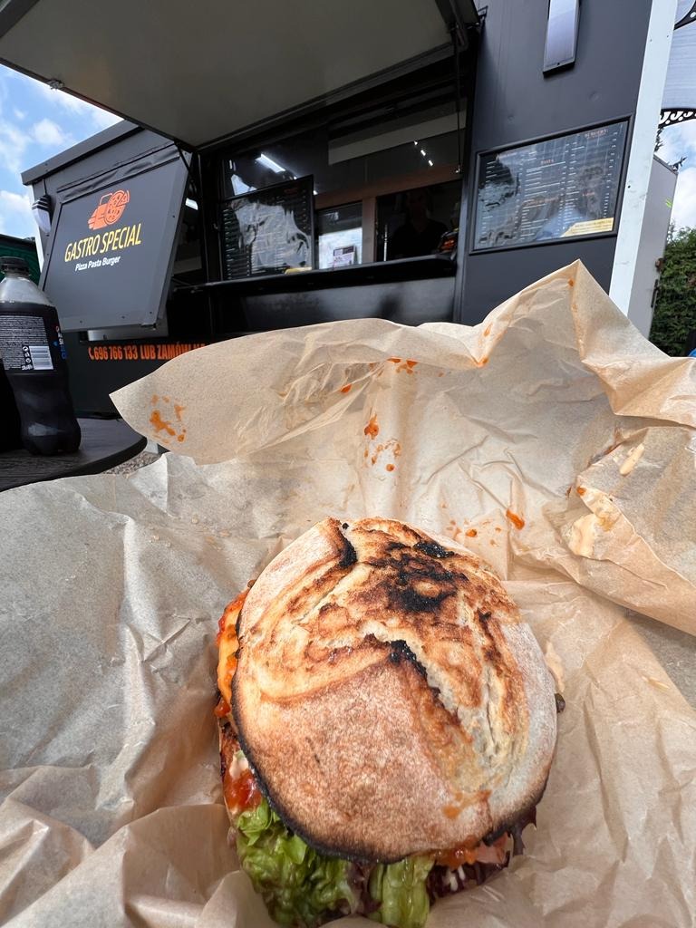 Burger z wołowiną i świeżą sałatą w rustykalnej bułce, podany w papierze na tle food trucka z logo 'Gastro Special'.