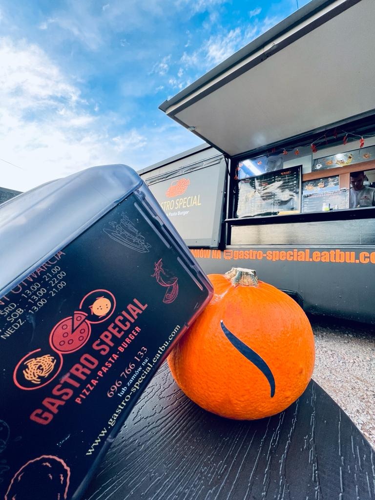 Pudełko z logo Gastro Special, dynia z wyciętym wzorem i food truck w tle na tle błękitnego nieba.
