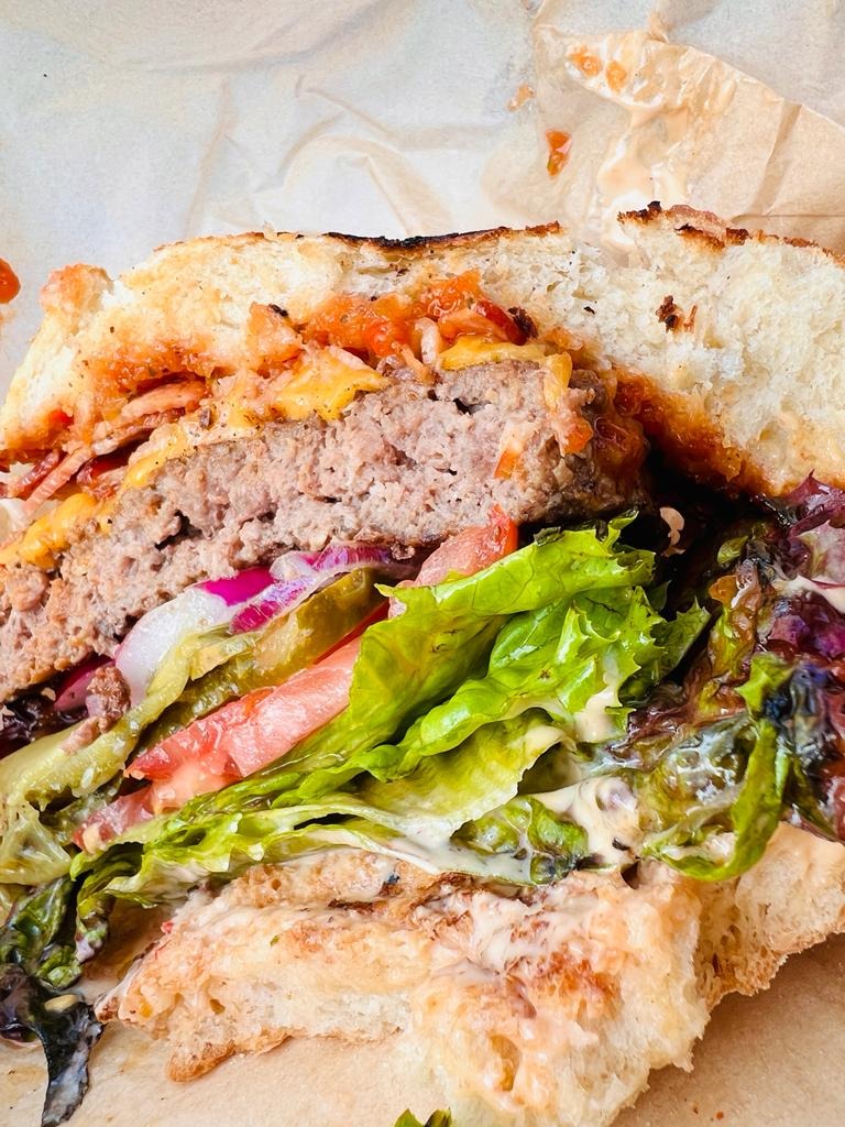 Przekrój burgera z wołowiną, serem cheddar, pomidorem, sałatą, piklami, czerwoną cebulą i sosem, ułożony na papierze.