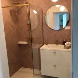 Nowoczesna łazienka z kabiną prysznicową typu walk-in, wykończona ceglastymi płytkami ułożonymi w jodełkę, złotą armaturą i okrągłym lustrem.