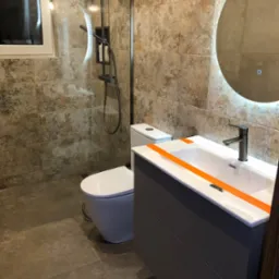 Nowoczesna łazienka z kabiną prysznicową typu walk-in, umywalką w szafce z zabezpieczeniem pomarańczową taśmą i okrągłym lustrem z podświetleniem LED.