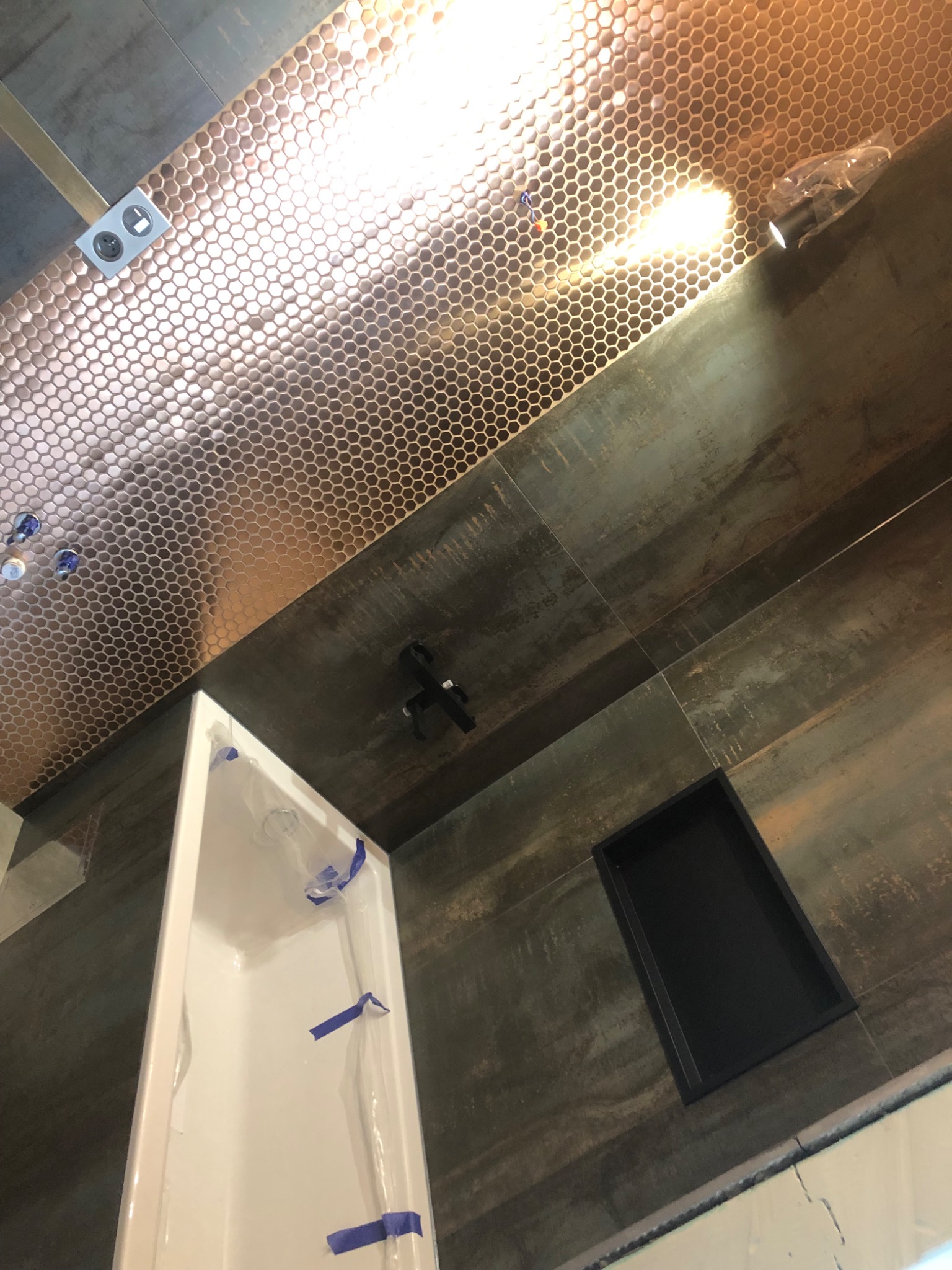 Narożnik łazienki w trakcie remontu: biała wanna zabezpieczona folią, ściany wyłożone ciemnymi płytkami imitującymi metal, sufit pokryty mozaiką w odcieniach miedzi.