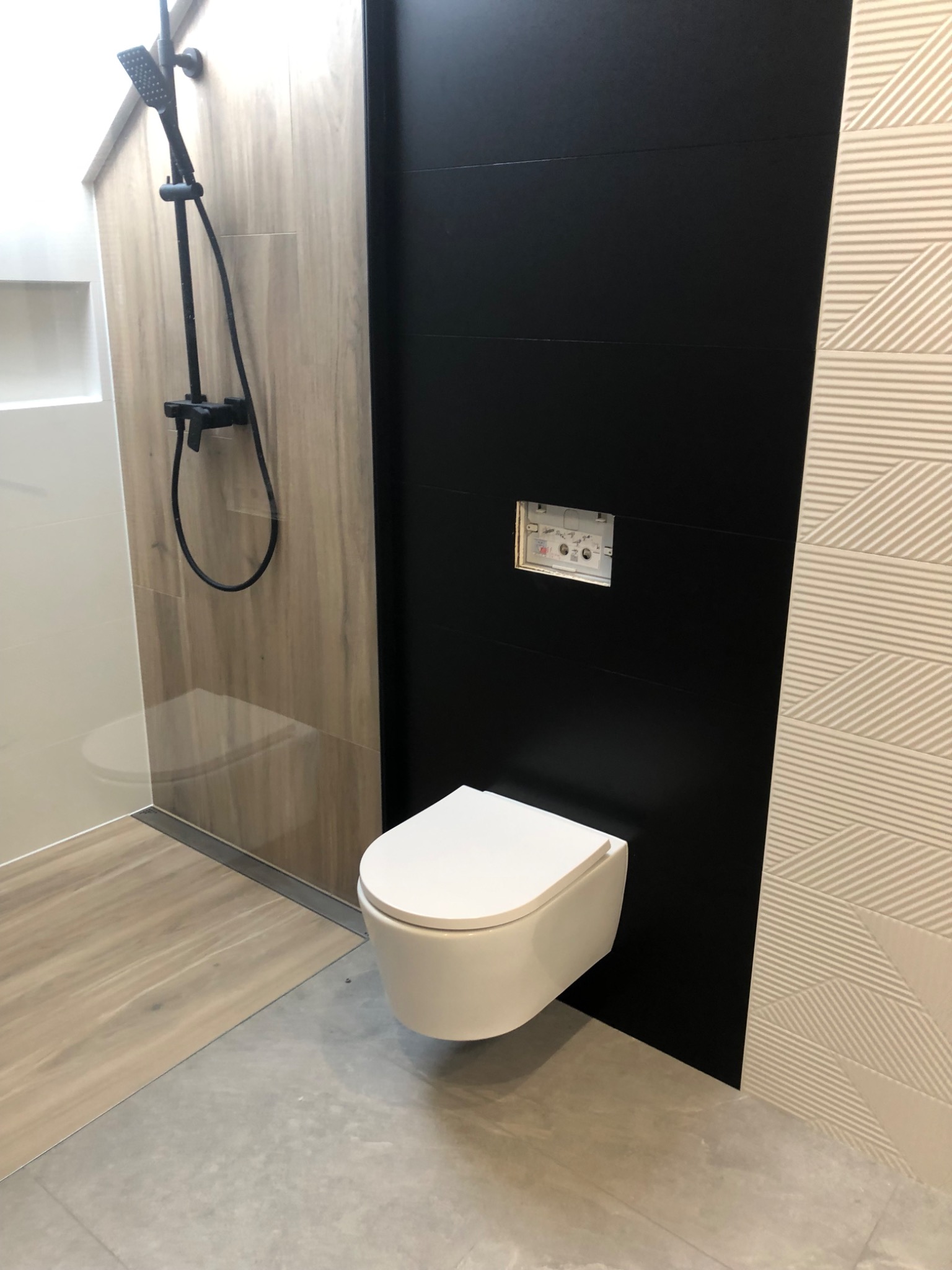 Nowoczesna łazienka z wiszącą toaletą, czarną ścianą i kabiną prysznicową z deszczownicą w kolorze czarnym, drewnopodobne i geometryczne płytki na ścianach.
