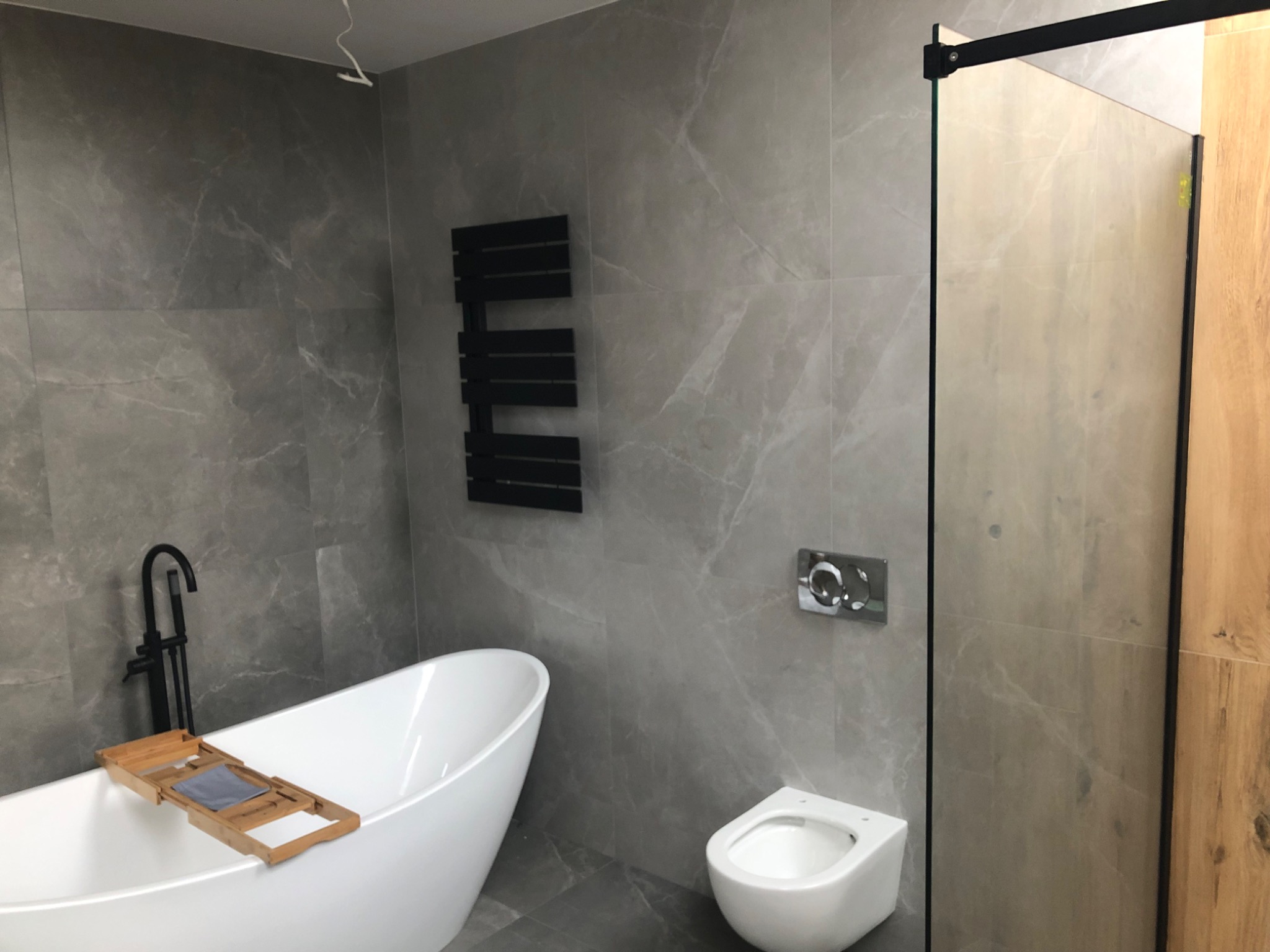 Nowoczesna łazienka z wanną wolnostojącą, czarną armaturą, grzejnikiem drabinkowym i płytkami imitującymi marmur w odcieniach szarości. Kabina prysznicowa z elementami drewnopodobnymi.