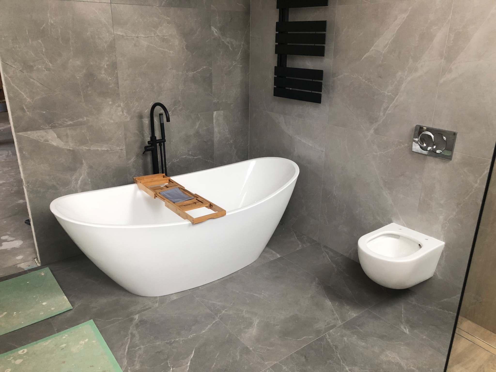 Nowoczesna łazienka z wolnostojącą białą wanną, czarną armaturą, wiszącą miską WC i szarą marmurową glazurą na ścianach, widoczne fragmenty niezakończonych prac wykończeniowych.