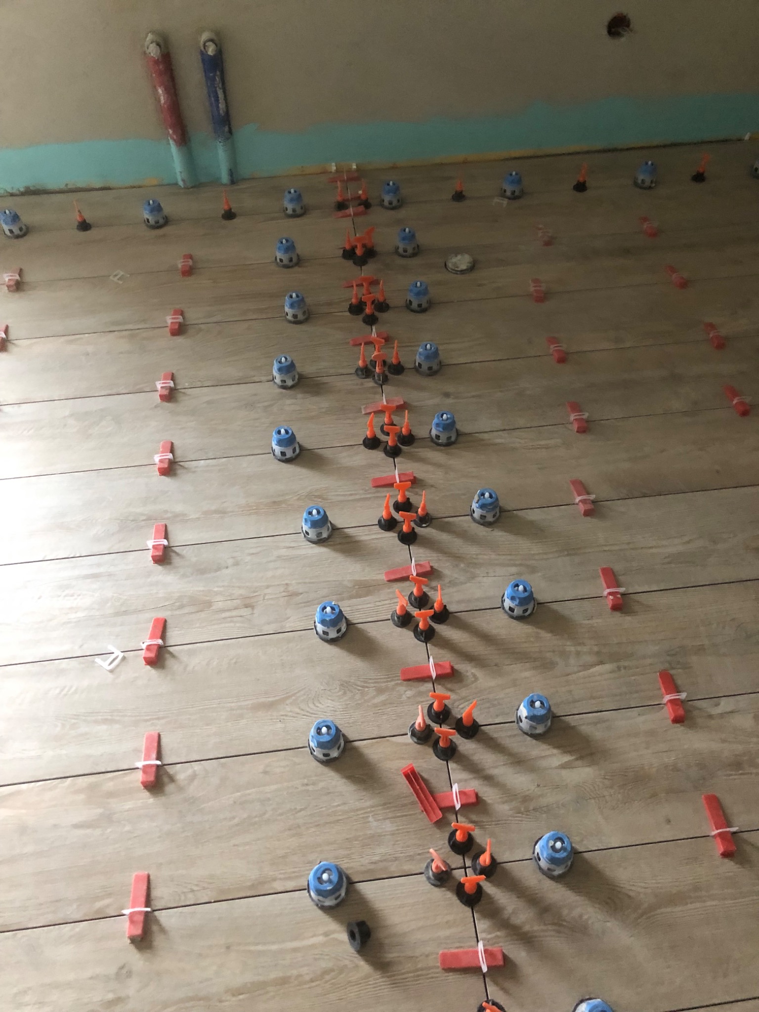 Układanie płytek podłogowych z użyciem systemu poziomowania: niebieskie klipsy i pomarańczowe kliny, widoczne rury instalacyjne przy ścianie.