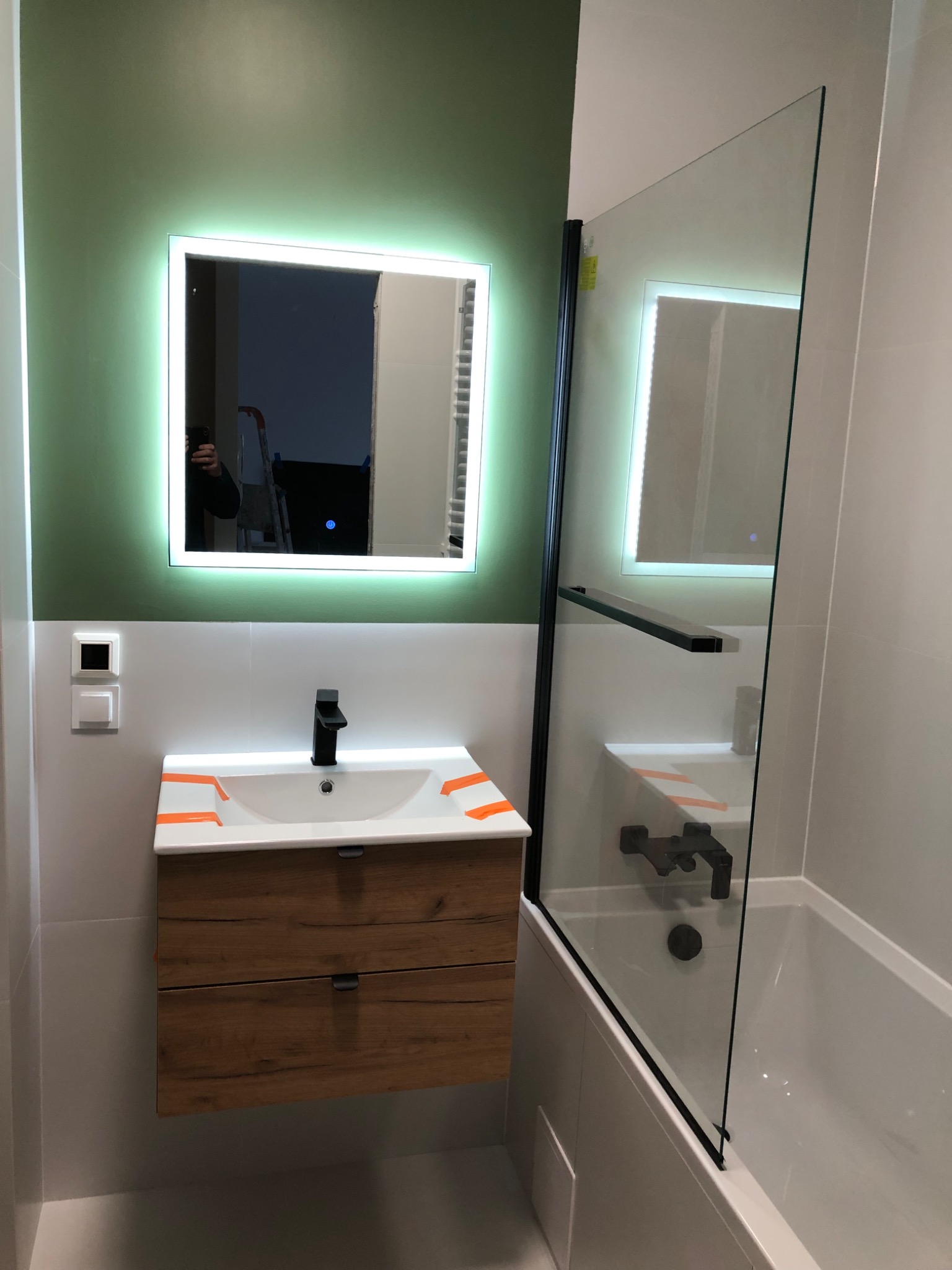 Nowoczesna łazienka z podświetlanym lustrem, umywalką z drewnianą szafką i szklanym parawanem nawannowym z czarnymi elementami.