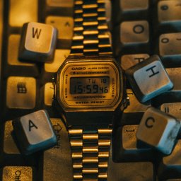 Jan Janota - Złoty zegarek Casio leżący na klawiaturze komputera, otoczony klawiszami z literami W, A, H, C i T, oświetlony ciepłym światłem.
