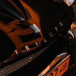 Jan Janota - Fragment czarnego, lśniącego samochodu Triumph z chromowanym napisem i pomarańczową tablicą rejestracyjną.