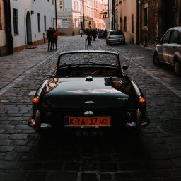 Jan Janota - Czarny kabriolet Triumph Spitfire zaparkowany na brukowanej uliczce w Krakowie, widok z tyłu, z odbiciami w karoserii i ludźmi w tle.