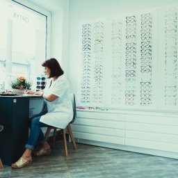 Jan Janota - Wnętrze salonu optycznego: kobieta w białym fartuchu siedzi przy biurku, obok ekspozycja okularów na ścianie, dekoracje roślinne, jasne światło wpadające przez okno.