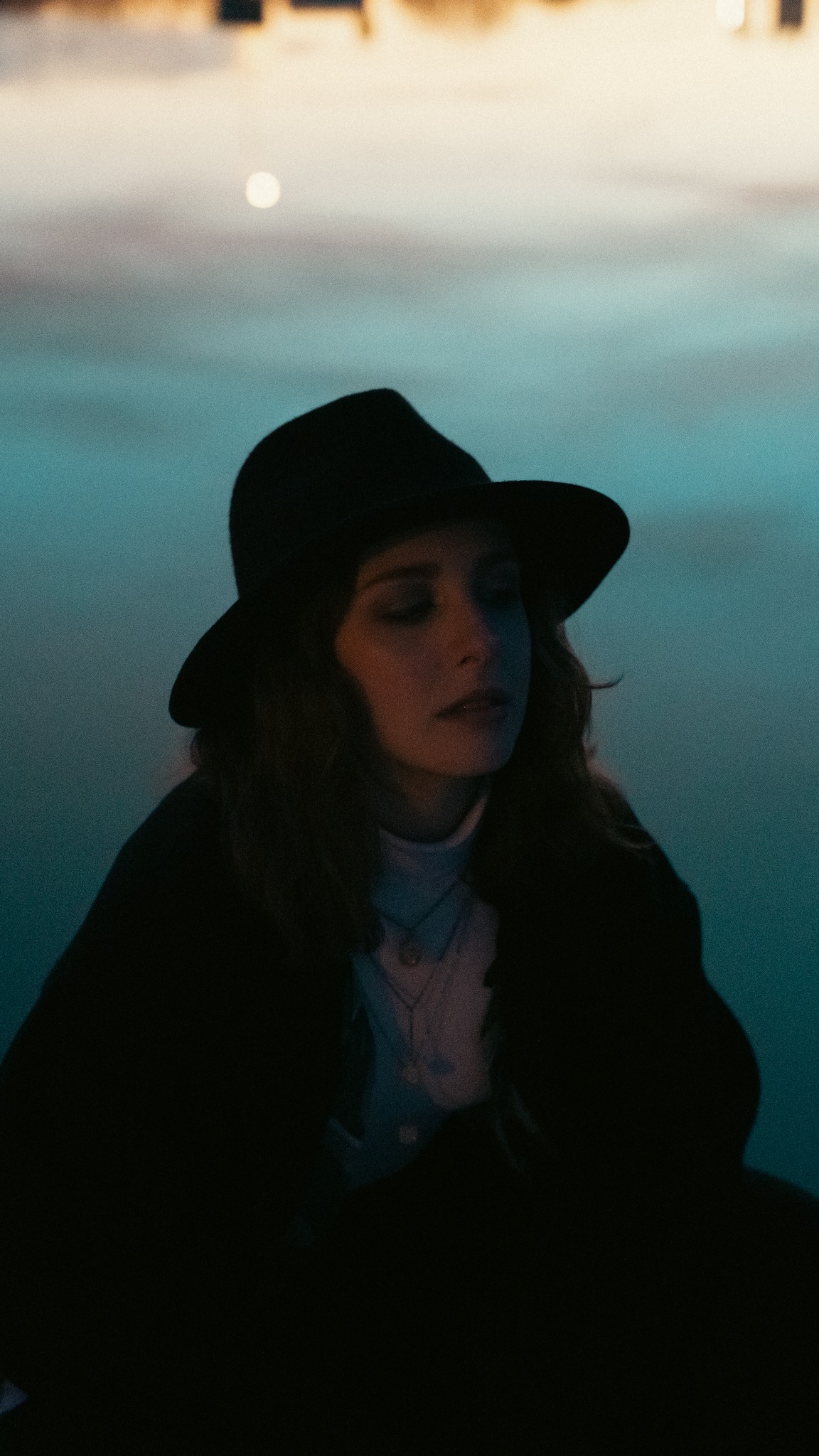 Portret kobiety w czarnym kapeluszu na tle rozmytego, niebieskiego tła z delikatnym światłem w górnym rogu.