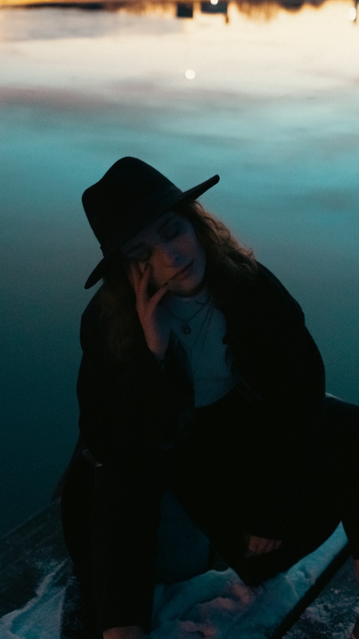 Zamyślona kobieta w czarnym kapeluszu i płaszczu, siedząca na tle jeziora o zmierzchu, z ręką opartą o twarz.