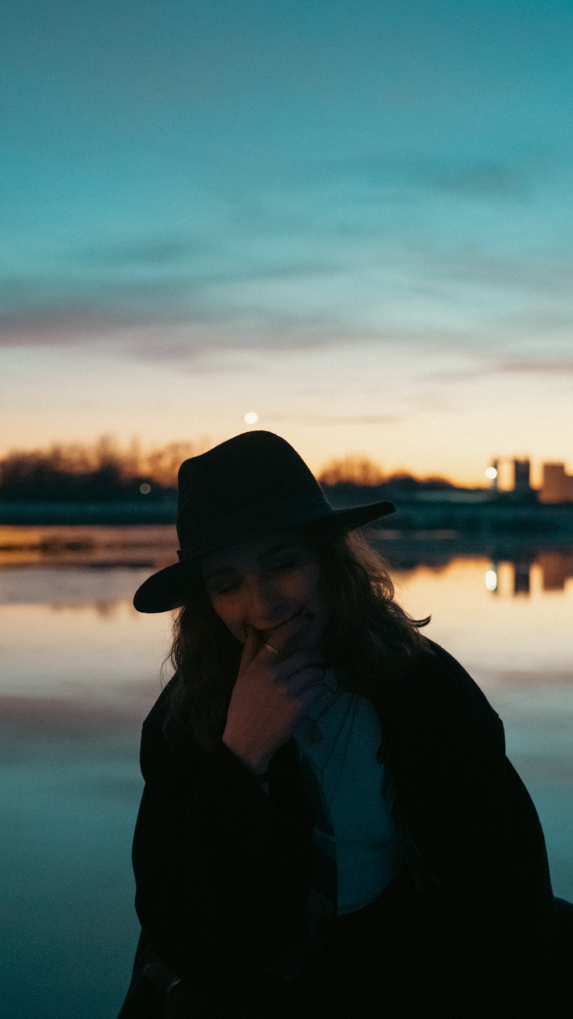 Portret kobiety w kapeluszu na tle zachodzącego słońca nad wodą, zarys budynków w oddali.