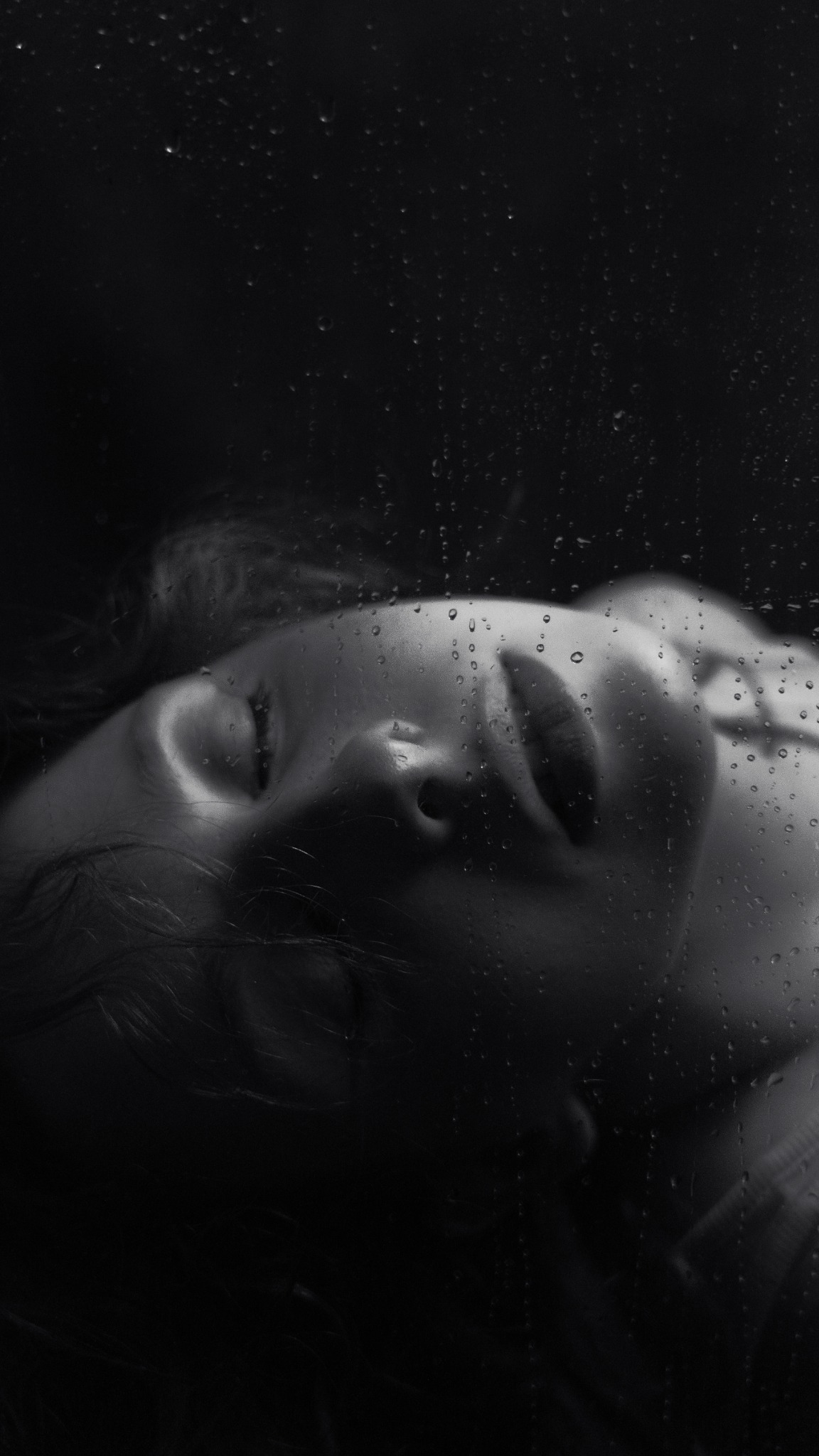 Monochromatyczne zbliżenie twarzy kobiety za szkłem pokrytym kroplami wody, z delikatnym oświetleniem podkreślającym rysy i tworzącym intymną atmosferę.