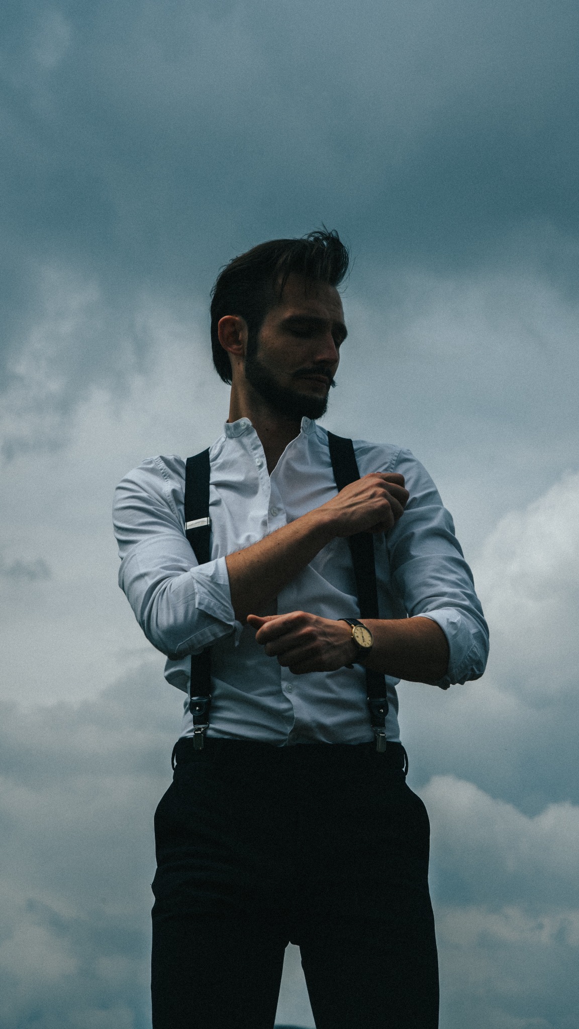 Mężczyzna w białej koszuli z szelkami, poprawiający mankiet na tle pochmurnego nieba, z widocznym złotym zegarkiem na ręce.