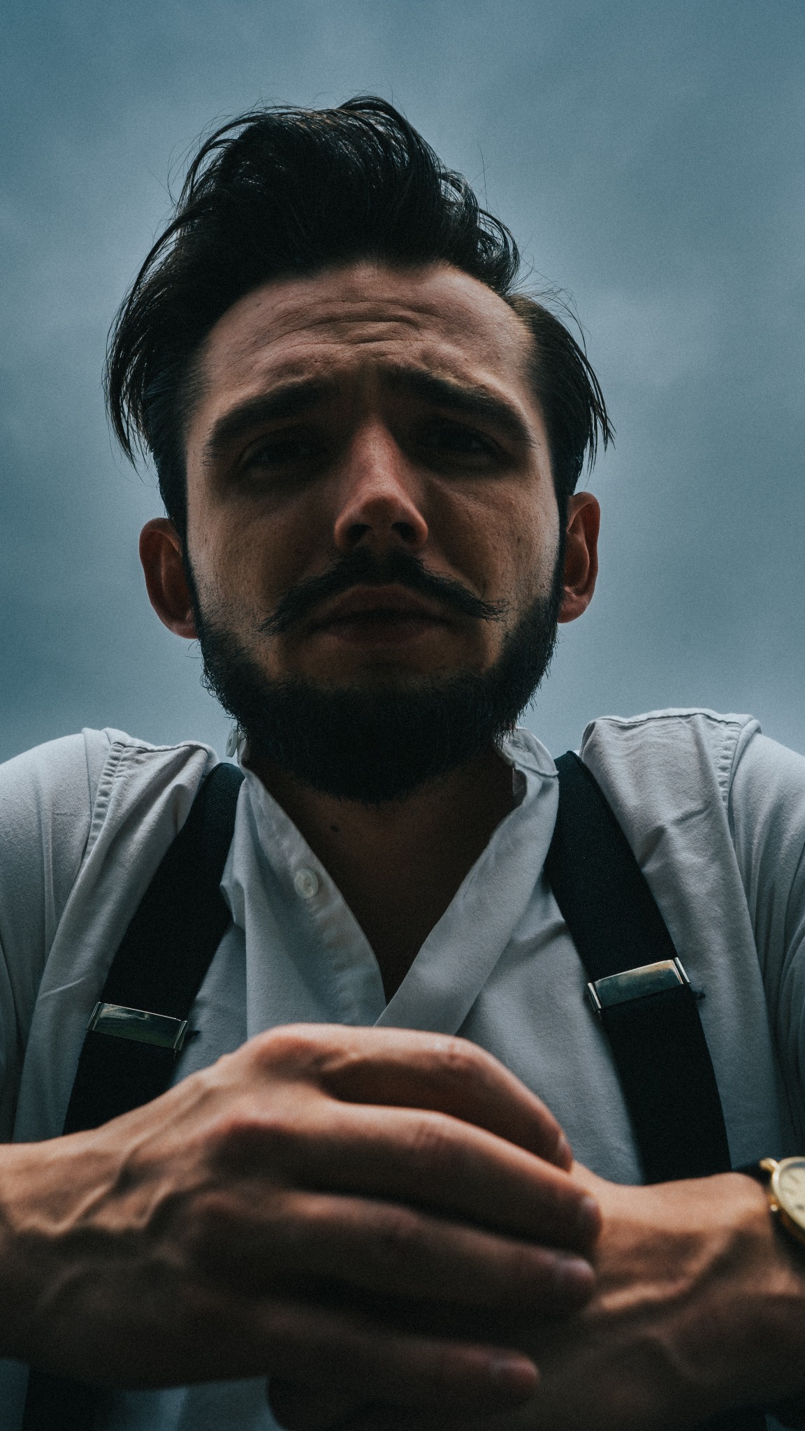 Portret mężczyzny z brodą i wąsami, ubrany w białą koszulę i szelki, spoglądający w dół z góry, z widocznym zegarkiem na nadgarstku i pochmurnym niebem w tle.