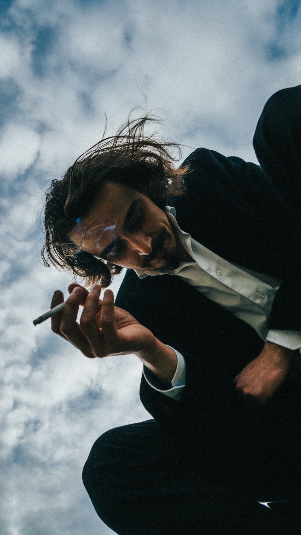 Mężczyzna w garniturze, widziany z dołu, trzyma zapalonego papierosa na tle pochmurnego nieba.