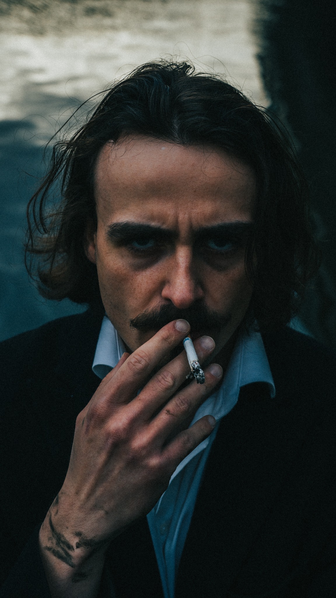 Portret mężczyzny z wąsami, palącego papierosa, w ciemnej marynarce i białej koszuli, z tatuażem na przedramieniu, uchwycony w klimatycznym, przyciemnionym oświetleniu.