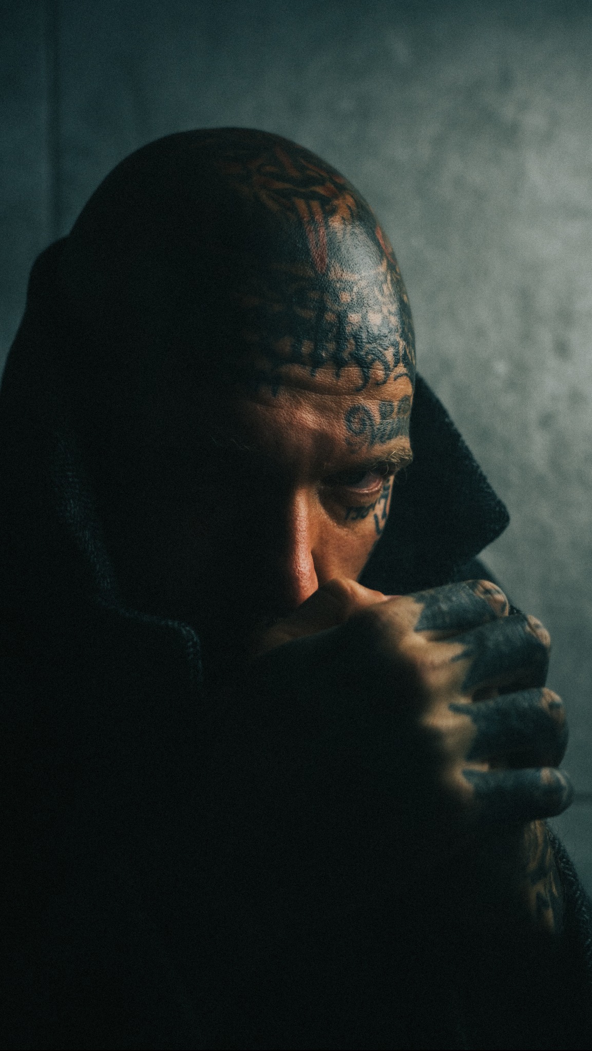 Portret mężczyzny z licznymi tatuażami na głowie i dłoniach, w ciemnym ubraniu, w melancholijnym nastroju, na tle szarej ściany.