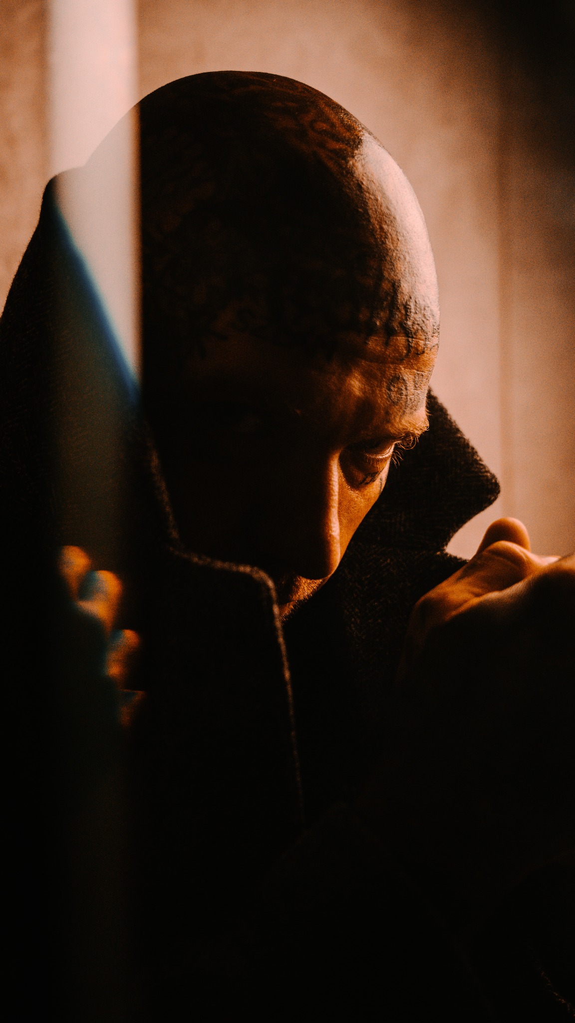 Mężczyzna w ciemnym płaszczu, z widocznymi tatuażami na głowie, spogląda w dół w dramatycznym oświetleniu, z elementami pomarańczowego światła.