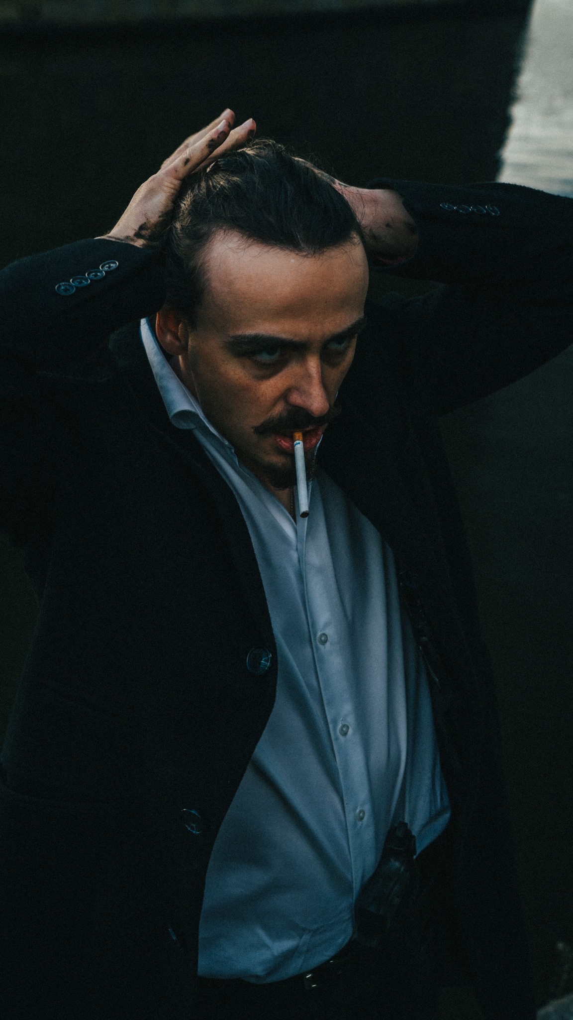 Portret mężczyzny z wąsami w rozpiętej koszuli i marynarce, trzymającego papierosa w ustach i brudnymi rękami poprawiającego włosy, z mrocznym spojrzeniem