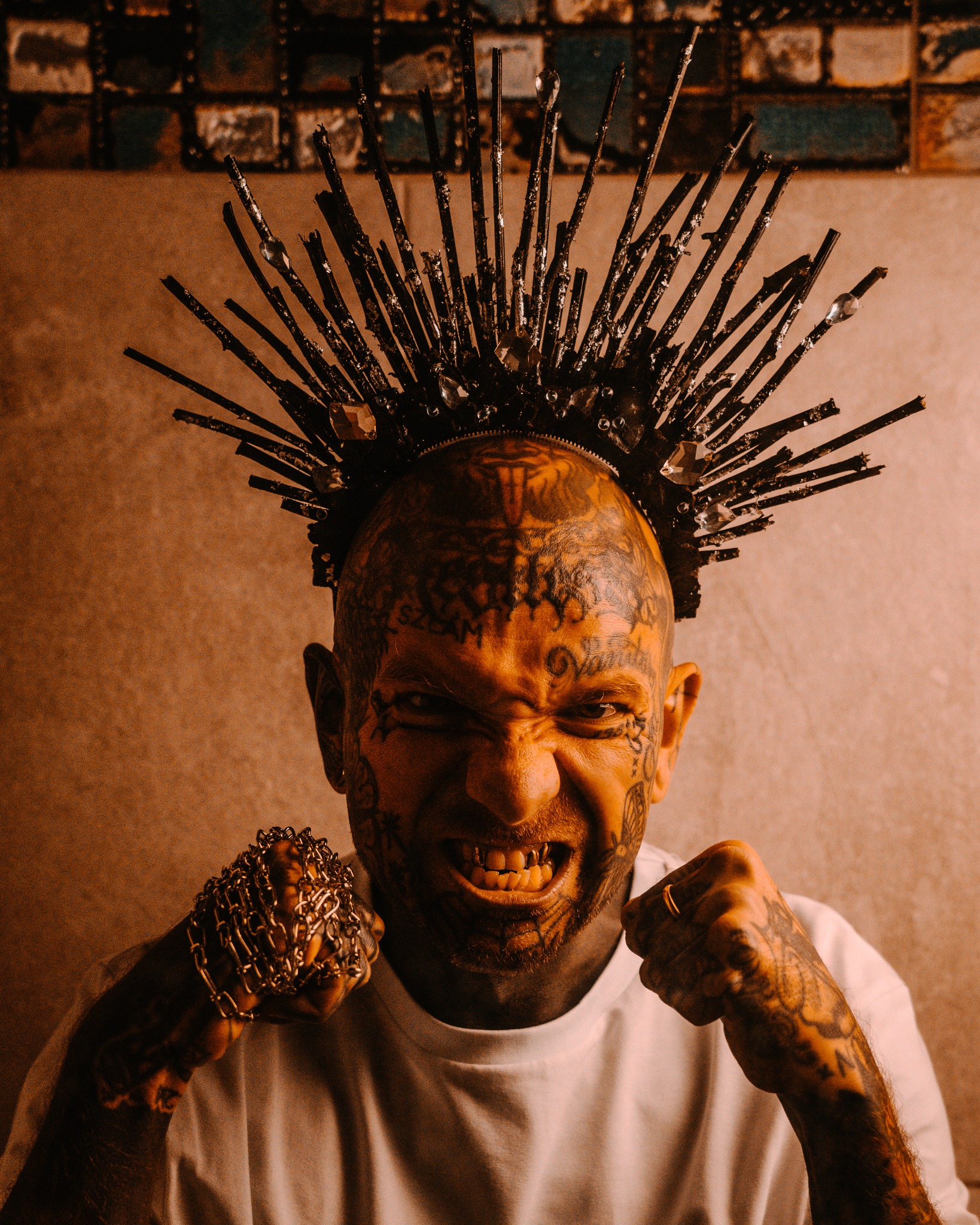Portret wytatuowanego mężczyzny z koroną z patyków i łańcucha, z grymasem na twarzy i złotymi nakładkami na zębach.