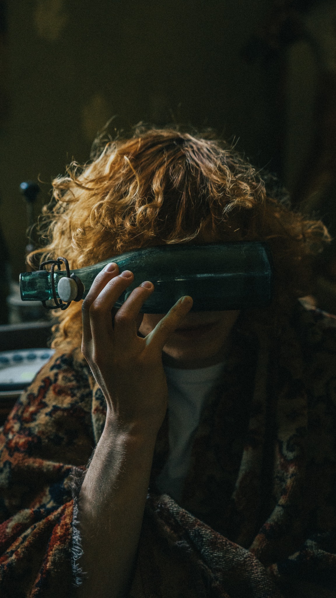 Osoba z rudymi kręconymi włosami trzyma przed twarzą starą, zieloną butelkę z zamknięciem na drucik i porcelanowym korkiem, częściowo zakrywając twarz, ubrana w wzorzystą narzutę.