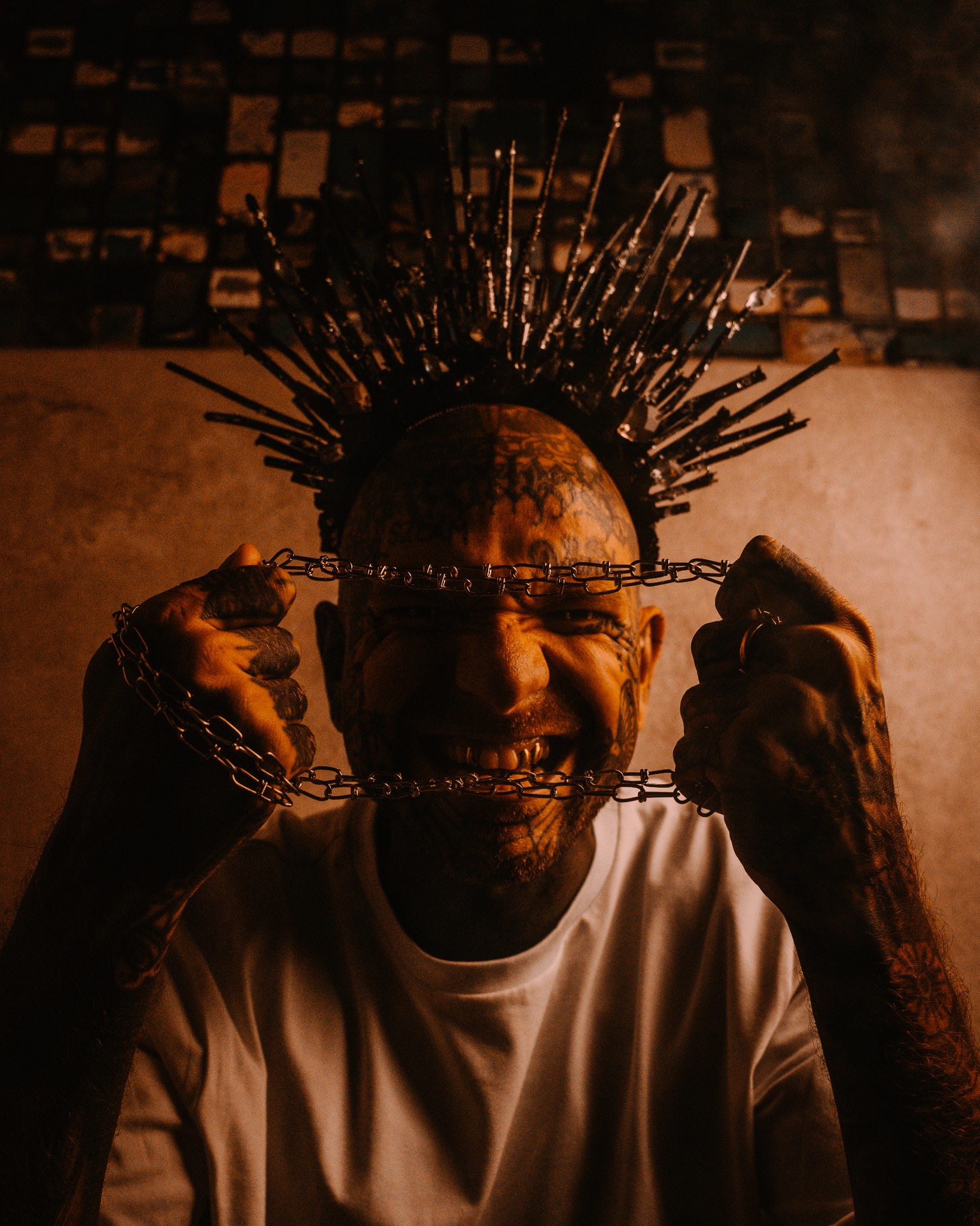 Portret mężczyzny z tatuażami na twarzy i koroną z kolców, uśmiechającego się przez łańcuch trzymany w dłoniach, z widocznymi złotymi nakładkami na zębach.