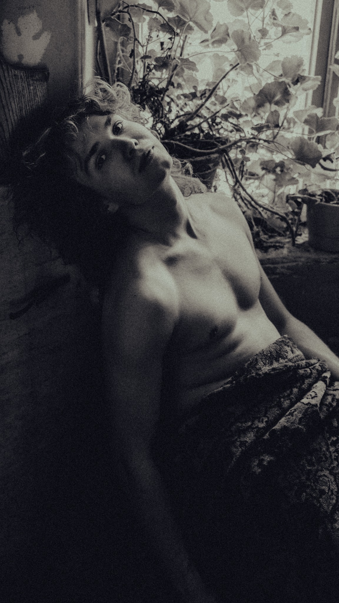 Mężczyzna z kręconymi włosami leży na tle okna z roślinami, w czarno-białej tonacji, z widocznym torsem i wzorzystym materiałem zakrywającym dolną część ciała.