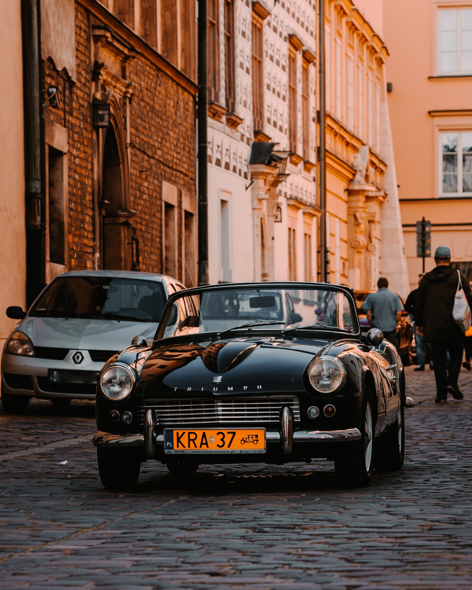 Czarny, klasyczny Triumph Spitfire zaparkowany na brukowanej ulicy w Krakowie, z widocznym numerem rejestracyjnym KRA 37, w tle budynki z cegły i piaskowca.