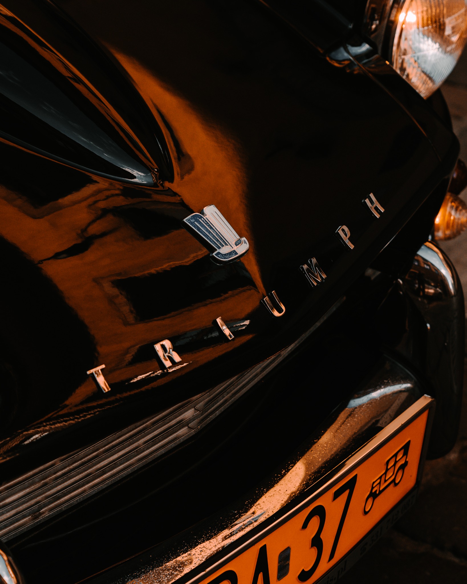Fragment czarnego, lśniącego samochodu Triumph z chromowanym napisem i pomarańczową tablicą rejestracyjną.