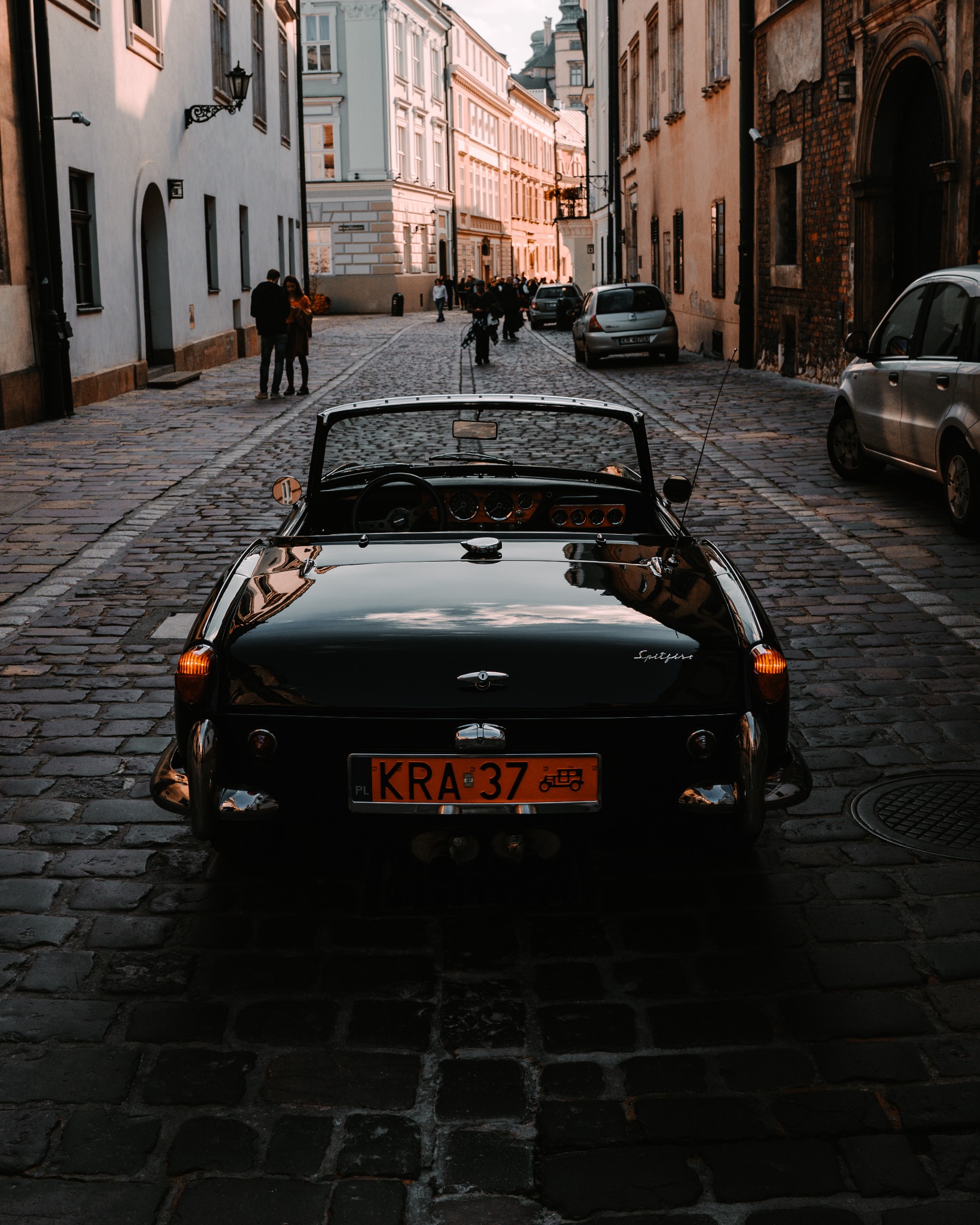 Czarny kabriolet Triumph Spitfire zaparkowany na brukowanej uliczce w Krakowie, widok z tyłu, z odbiciami w karoserii i ludźmi w tle.