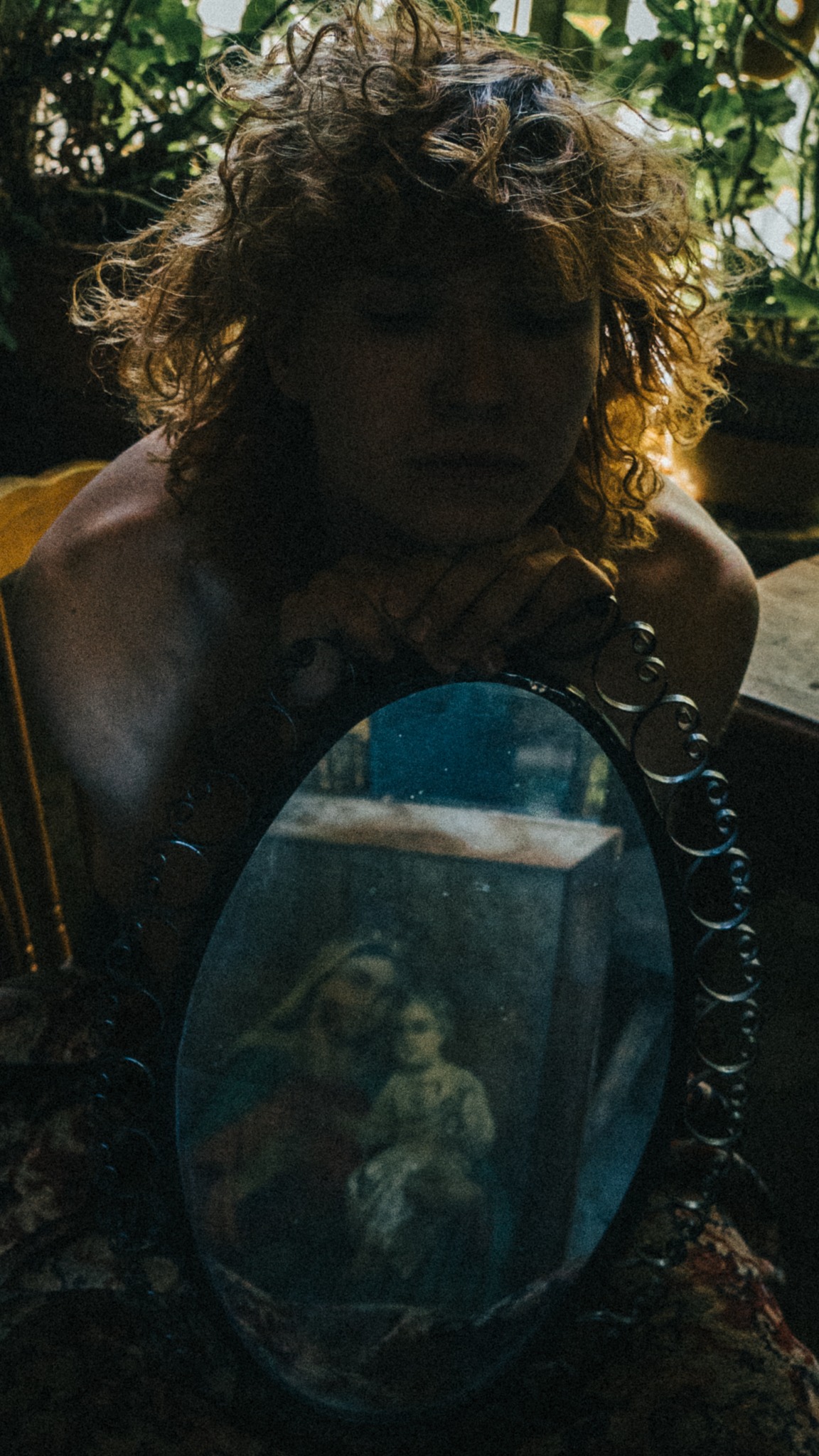 Portret kobiety z kręconymi włosami, opierającej brodę na dłoniach, z owalnym lustrem z ornamentami, w którym odbija się obraz Matki Boskiej z Dzieciątkiem, w tle rośliny doniczkowe.