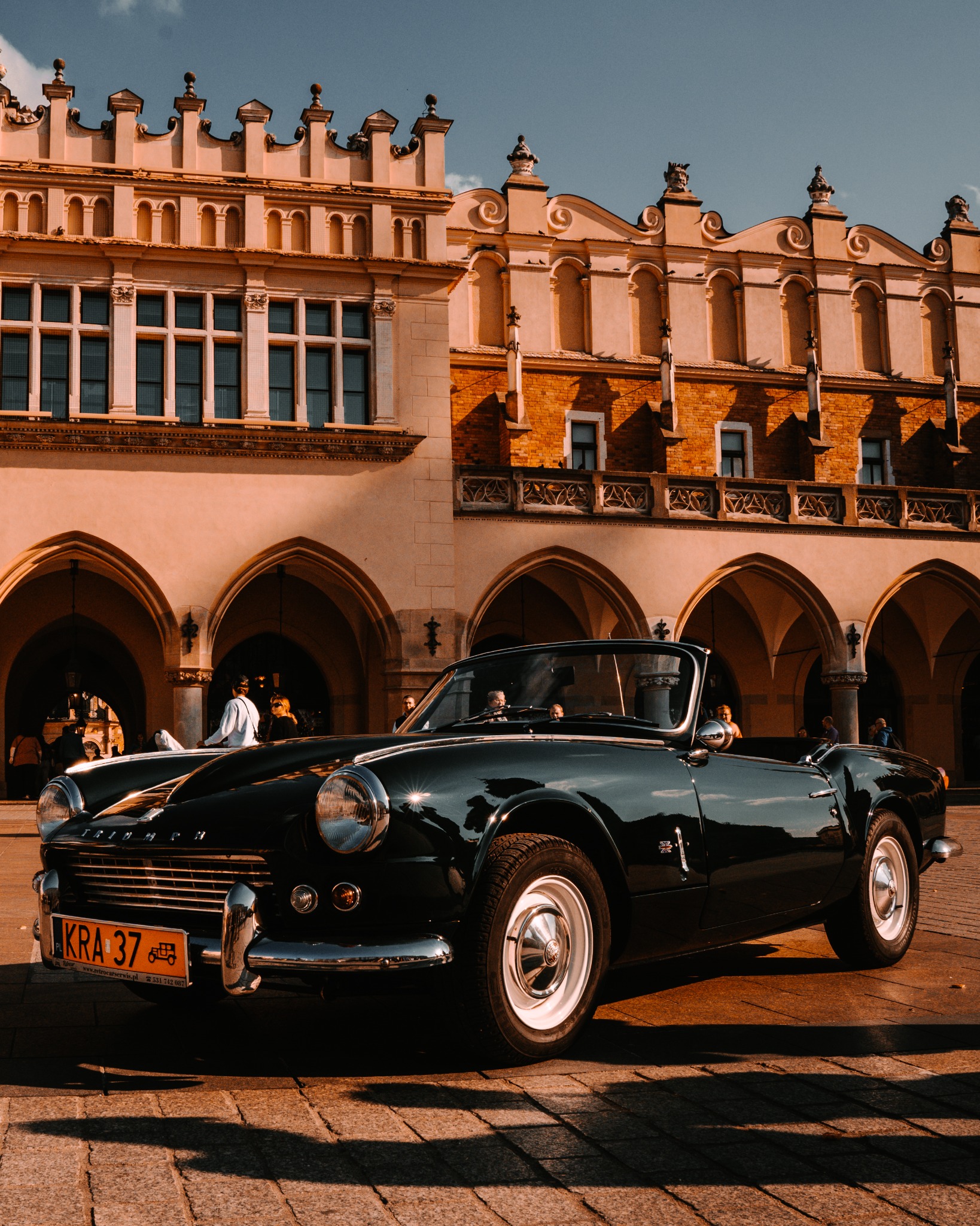 Czarny kabriolet Triumph Spitfire z tablicą rejestracyjną KRA 37 na tle Sukiennic w Krakowie, sfotografowany w ciepłym świetle.