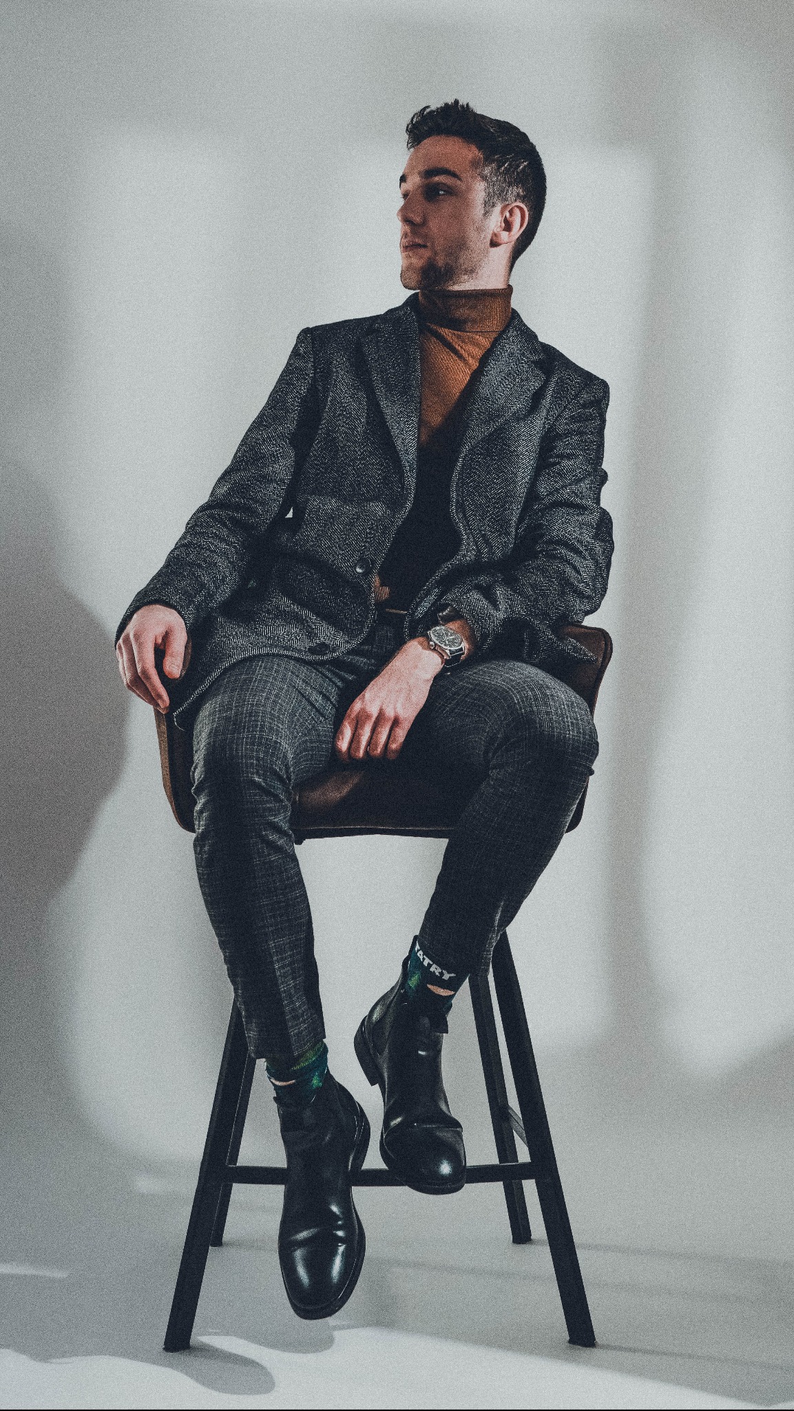 Mężczyzna w tweedowym garniturze i golfie pozuje siedząc na krześle w studio w Krakowie, prezentując stylizację z eleganckim zegarkiem i nietypowymi skarpetkami z napisem.