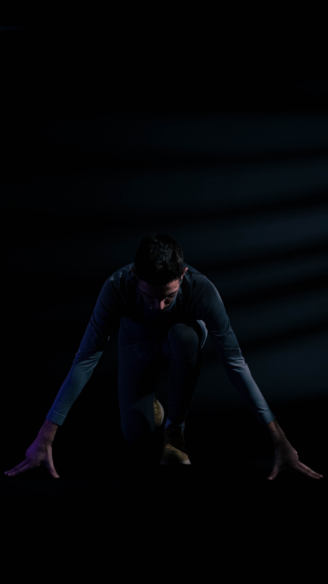 Mężczyzna w pozycji startowej na ciemnym tle, oświetlony z góry, gotowy do biegu, z rękami wyciągniętymi do przodu.