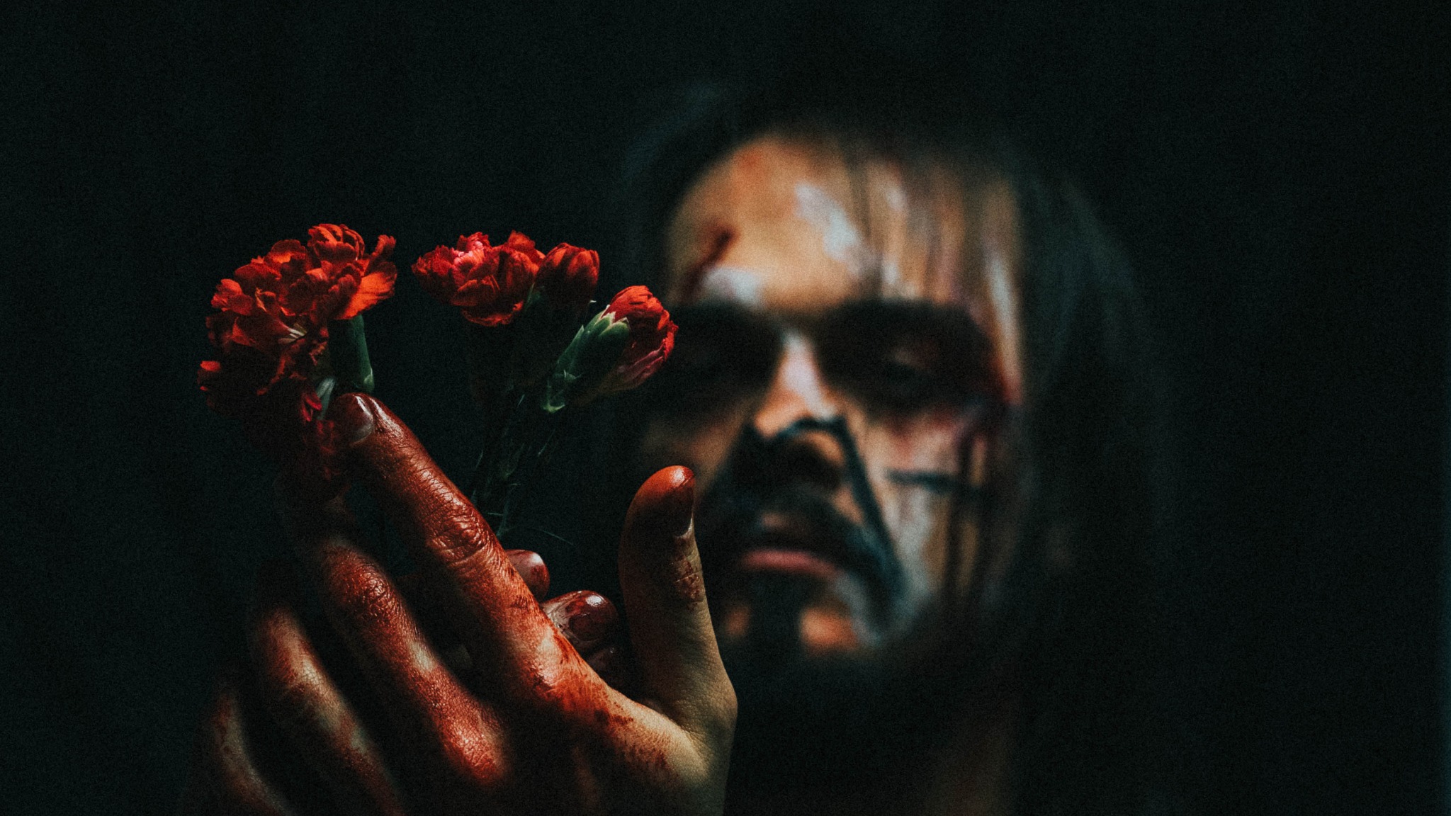 Mężczyzna z pomalowaną twarzą trzyma w zakrwawionej dłoni bukiet czerwonych goździków na ciemnym tle.