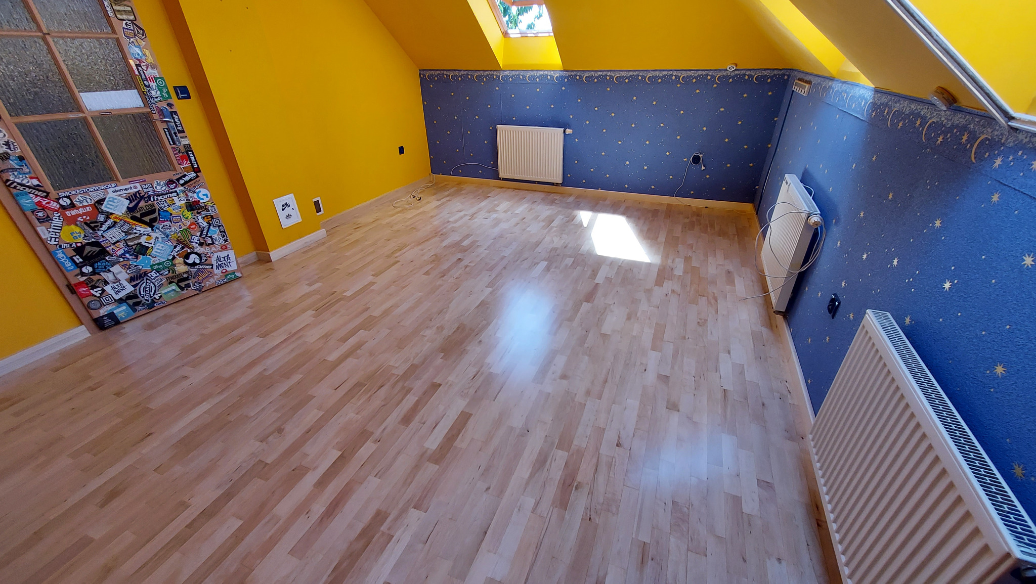 Jasny, nowo ułożony parkiet w pokoju na poddaszu z żółtymi i niebieskimi ścianami, okno dachowe i drzwi oklejone naklejkami.
