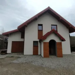 Dwukondygnacyjny dom z białą elewacją i brązowym dachem, widok z zewnątrz na szarym tle nieba, z elementami dekoracyjnymi imitującymi drewno przy wejściu i oknach.