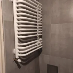 Biały, drabinkowy grzejnik łazienkowy zamontowany na szarej, płytkowej ścianie. Widoczne przyłącze z zaworem termostatycznym i rewizja kanalizacyjna w ścianie.