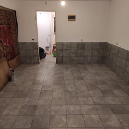 Arkadiusz Oklej - Układanie szarych, kwadratowych płytek na podłodze i częściowo na ścianach w pomieszczeniu. W tle widoczna osoba w trakcie pracy oraz pozostałości po remoncie.