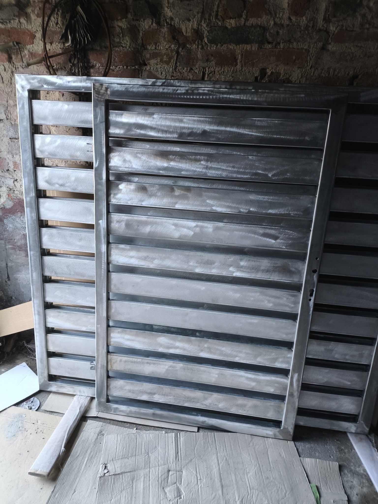 Dwa metalowe, surowe przęsła ogrodzeniowe z poziomymi lamelami, o szlifowanej powierzchni, oparte o ceglaną ścianę i karton. Widoczny surowy stan przed malowaniem.