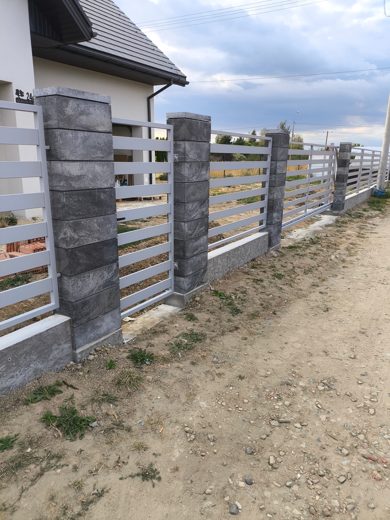 Nowoczesne ogrodzenie panelowe z szarymi słupkami z bloczków betonowych, na tle domu z szarą dachówką i pochmurnego nieba. Widok z zewnątrz posesji.