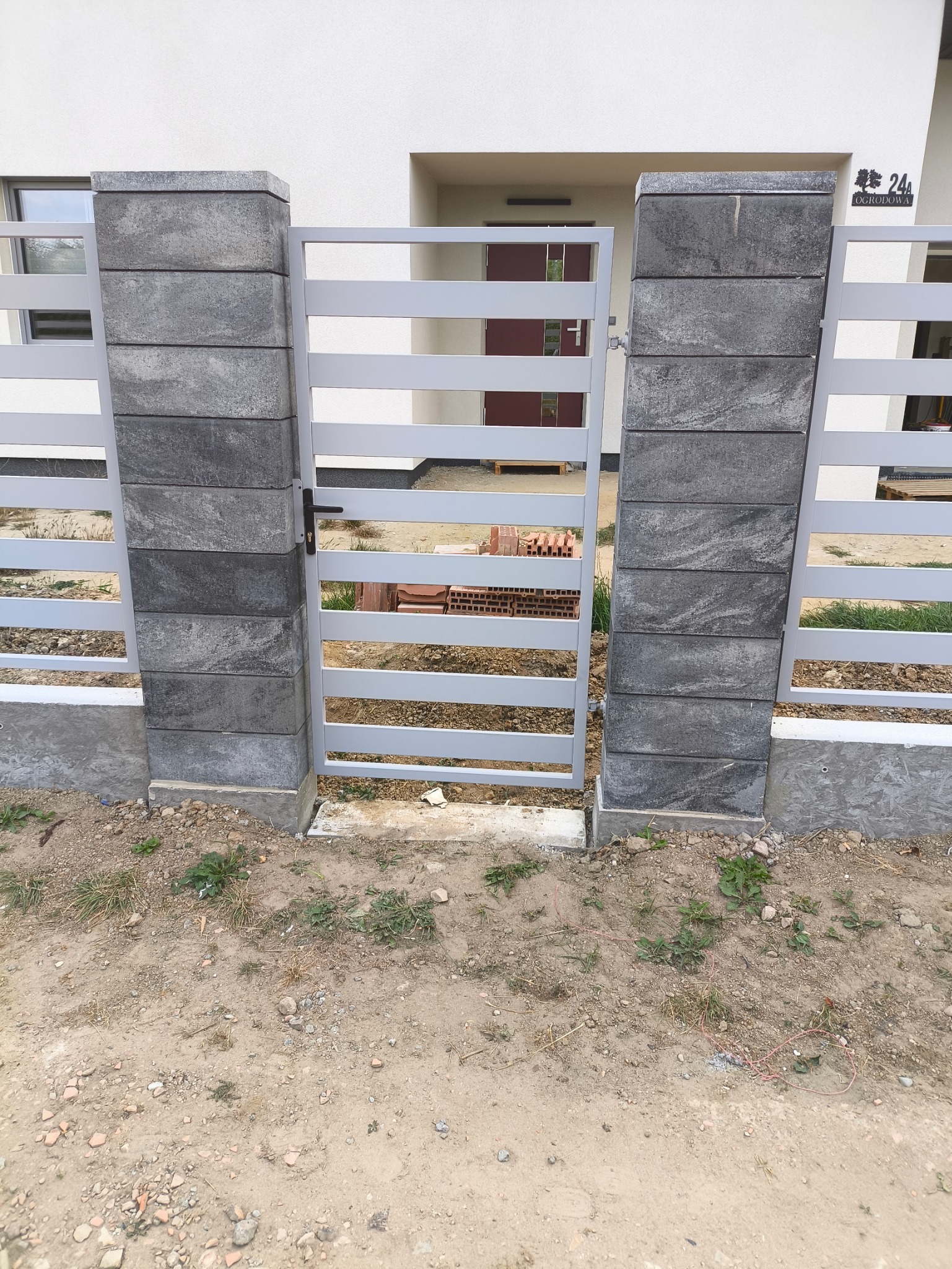 Nowoczesna, szara furtka z poziomych płaskowników, osadzona między dwoma słupkami z szarej kostki. W tle widoczny budynek z białymi ścianami i brązowymi drzwiami.