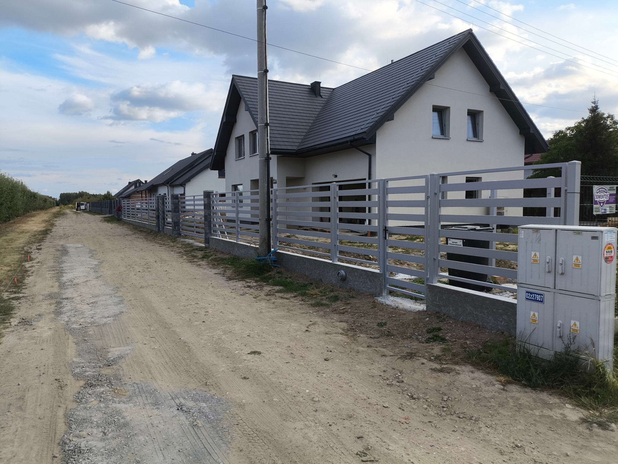 Szara, pozioma, metalowa palisada ogrodzeniowa przed szeregiem domów jednorodzinnych. Widok na drogę gruntową i pochmurne niebo.